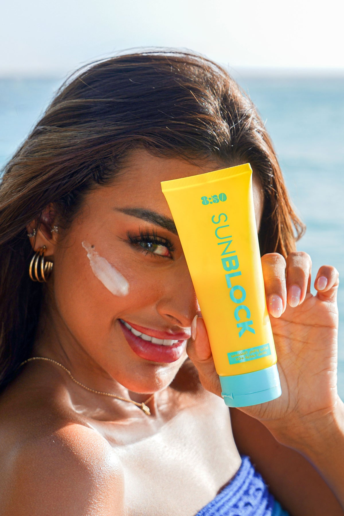 Model holding 8:50 Sunblock SPF 50 by the sea, cream on her cheek